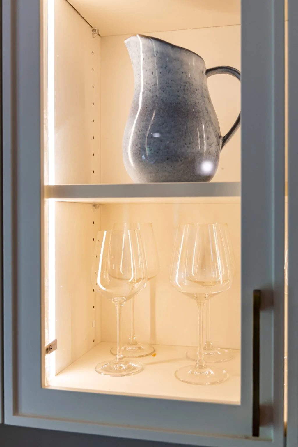 Lit display niche holding a metal pitcher and two wine glasses on oak shelving.