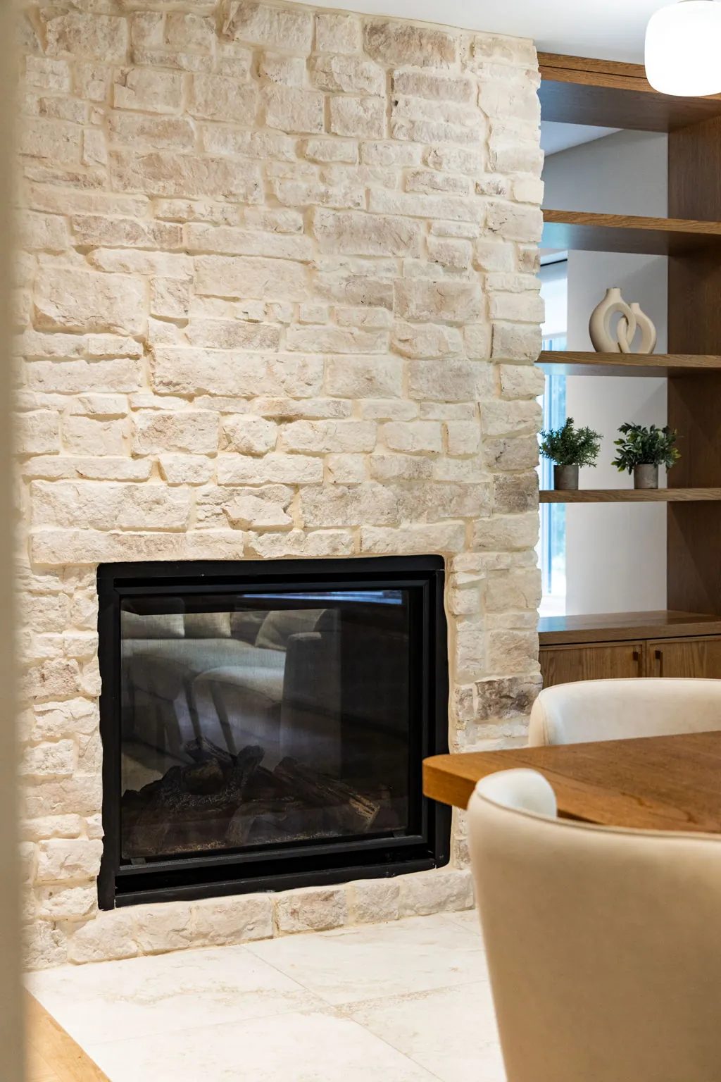 A black glass-fronted fireplace is set into pale rough-cut stone beside open oak shelving and a wood dining table with cream chairs.