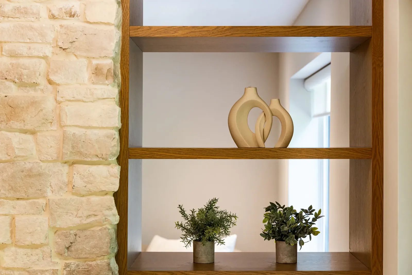 Two cream looped ceramic vessels and two small potted plants sit on open oak shelves beside a pale stone fireplace.