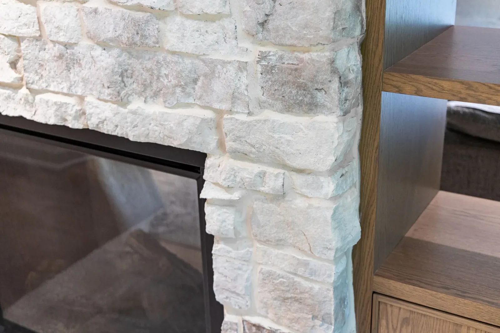 A close view shows the junction of pale rough-cut fireplace stone, a black metal firebox frame and open oak shelving.