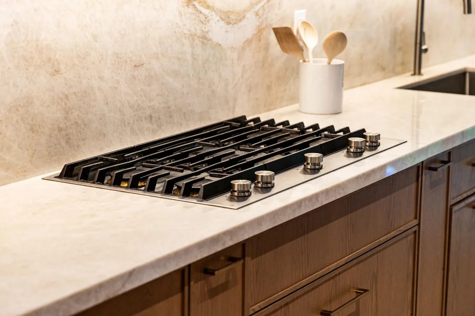A black gas cooktop with brushed metal knobs is inset into a pale stone counter above dark oak drawers, with a utensil crock near the sink.