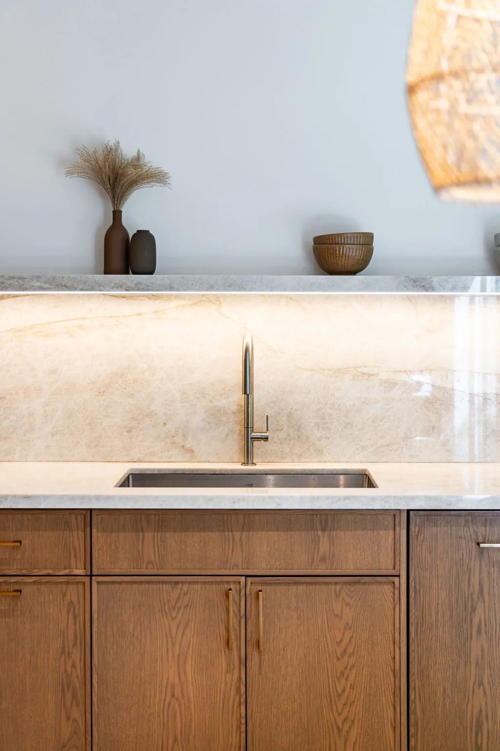 A stainless steel sink and slim faucet are set into a pale stone counter above oak cabinets, beneath an illuminated stone backsplash and a ledge of brown vessels.