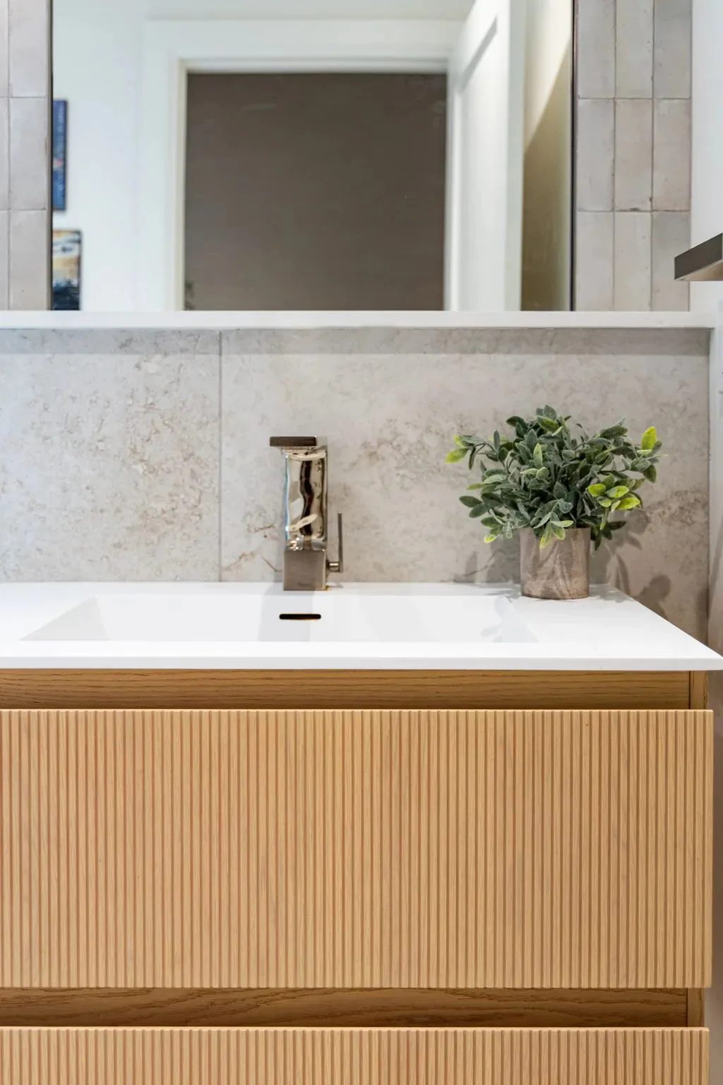 A bathroom vanity with a fluted pale wood front, white rectangular sink, chrome faucet and small potted plant stands below a wide mirror, with the bathroom type not visible.