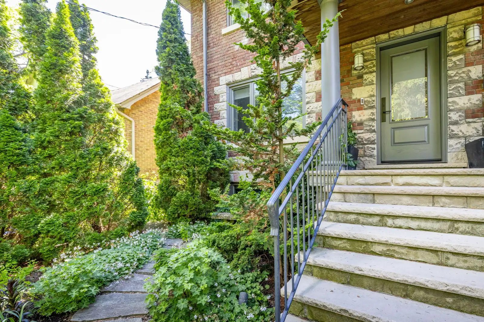 Brick and stone exterior with landscaped stone steps, an iron railing, and a sage front door set among mature greenery.