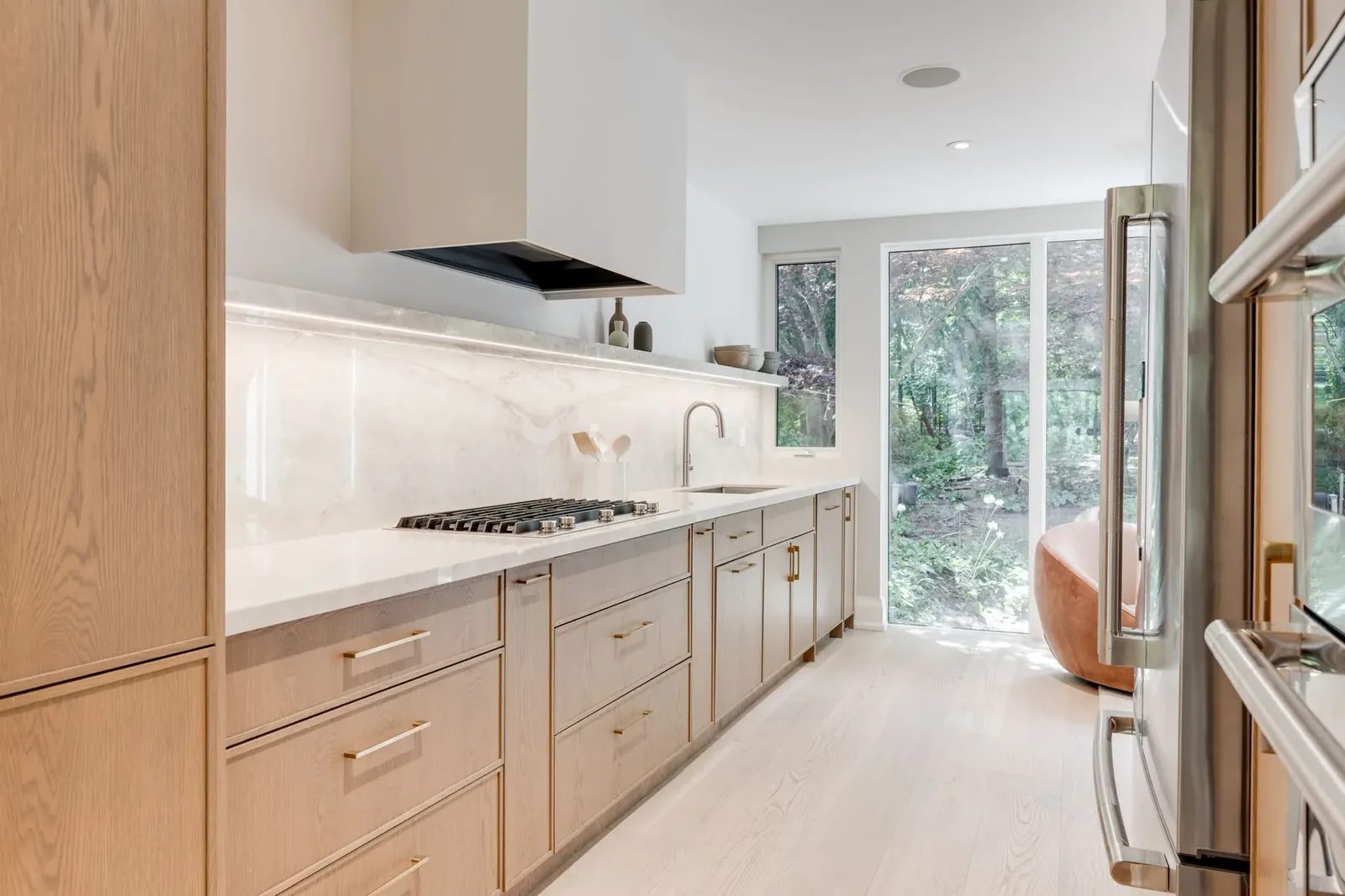 Kitchen run beneath a white plaster hood, with a marble slab backsplash and lit stone shelf, ending at full-height glass to the wooded garden.