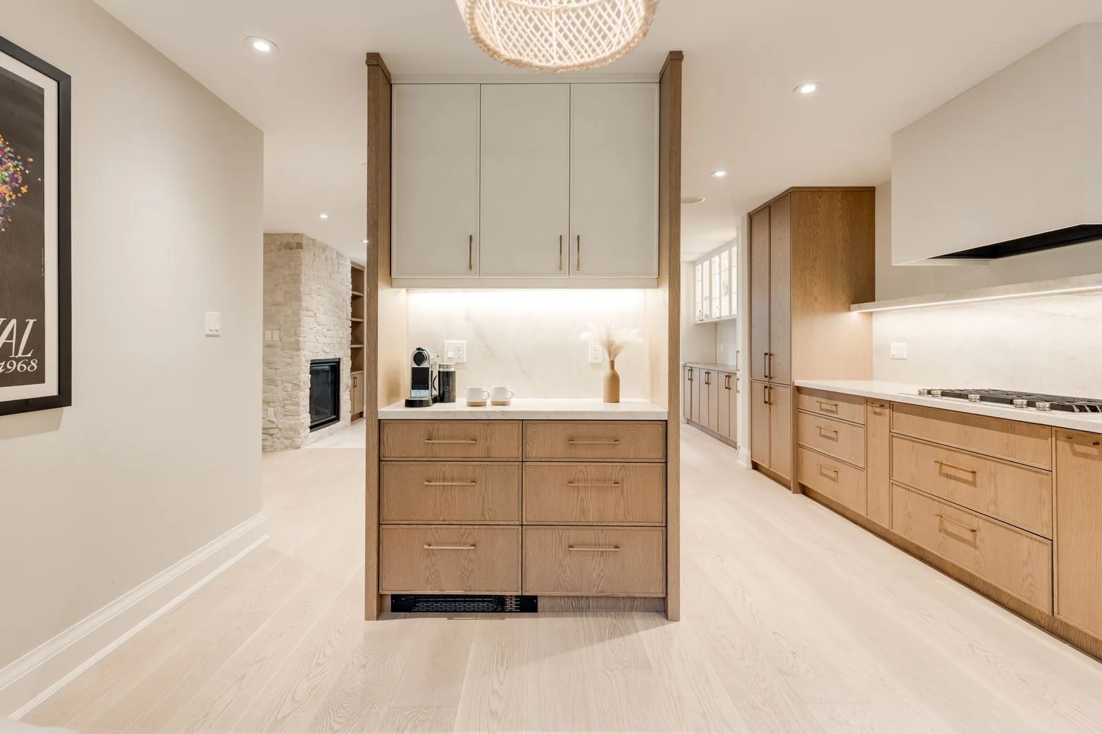White oak servery peninsula with a coffee station beneath a woven pendant, looking through to the kitchen run and the stone fireplace beyond.