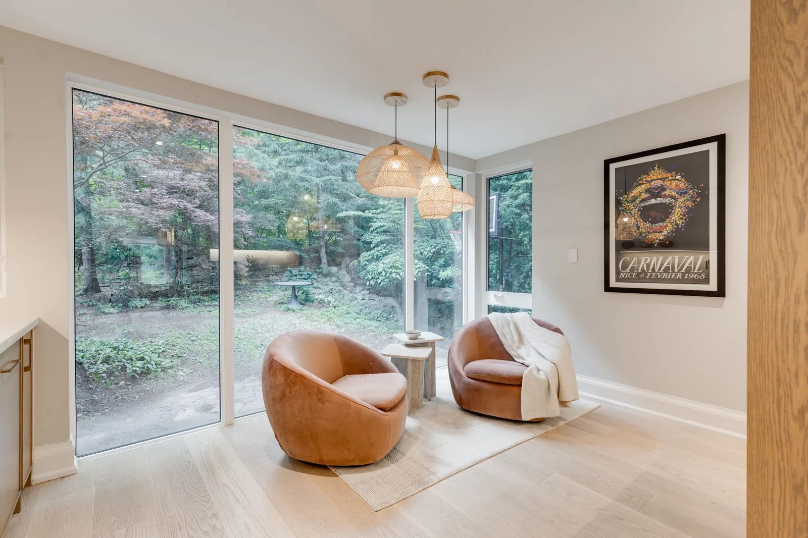 Sitting area with two orange leather swivel chairs and a stone side table beneath cluster pendants, beside a wall of windows onto the wooded ravine.