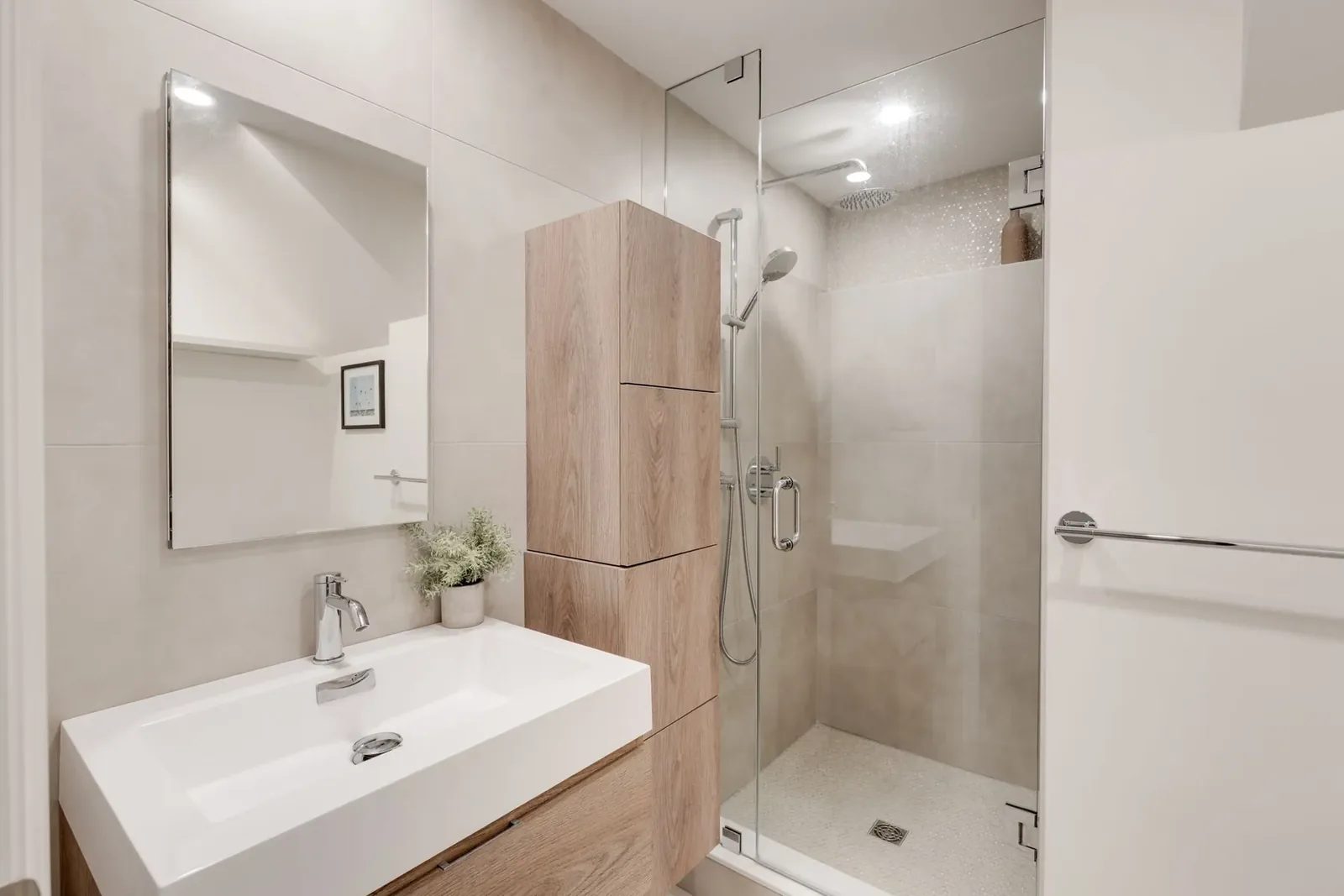 Bathroom with a wide white basin and chrome faucet, a wood-grain storage tower, and a frameless glass shower with a round rainfall head.