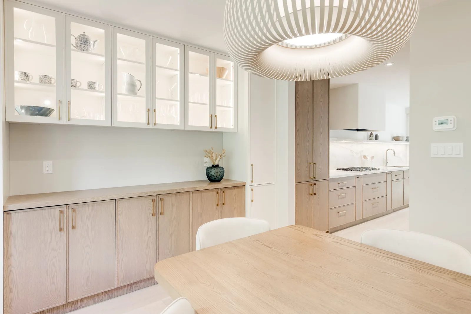 Dining room with an oak table under a slatted pendant, backed by a servery of glass-front upper cabinets over white oak drawers.