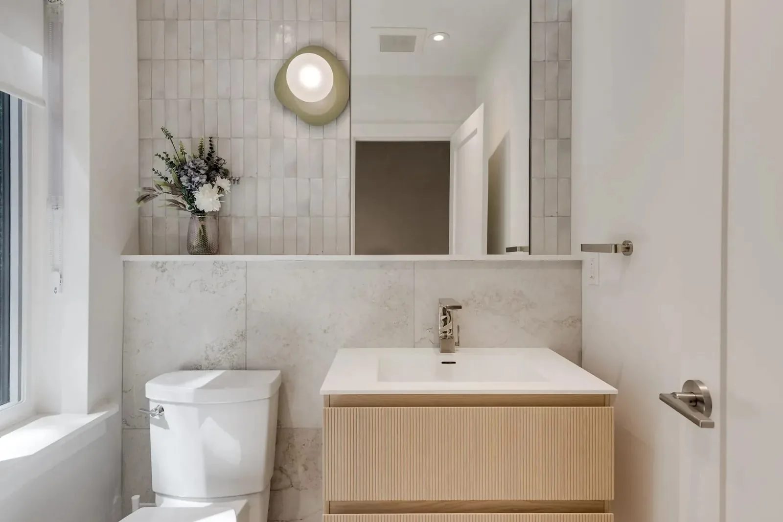 Powder room with a fluted white oak floating vanity, a stacked vertical-tile feature wall over stone-look wainscot, an olive wall sconce, and a wide mirror.