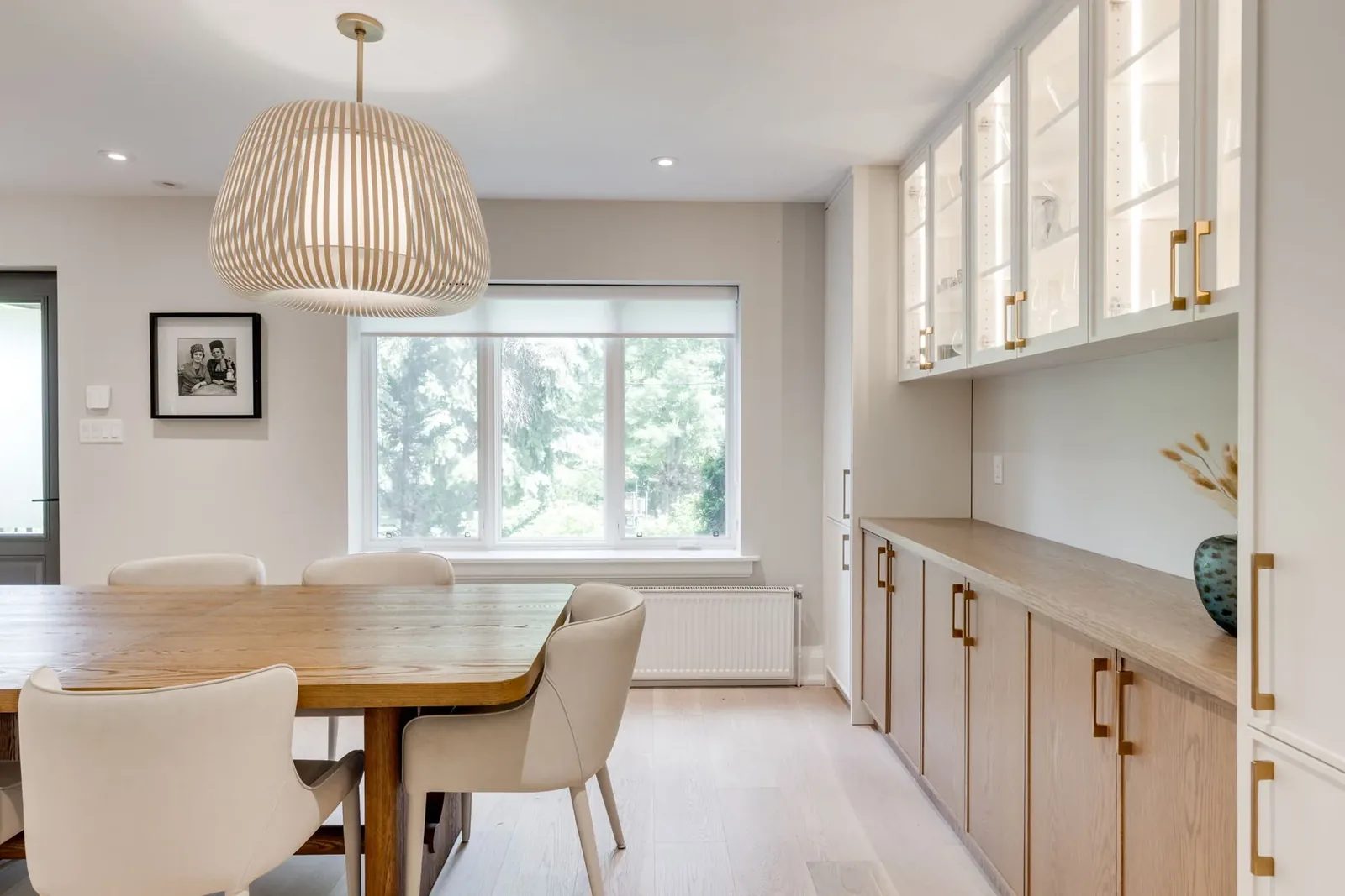 Dining room servery run of lit glass-front uppers and white oak lowers with brass pulls, beside the table and a shaded picture window.