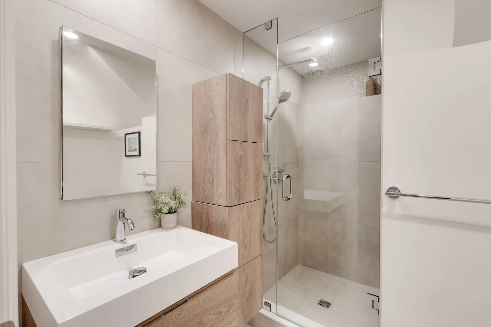 Bathroom with a frameless glass walk-in shower, a white oak storage tower, and a long white trough sink on a floating vanity.