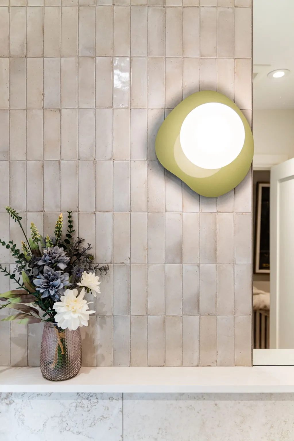 Lit olive sconce on the stacked cream tile of the powder room, above a textured glass vase of flowers on the stone ledge.