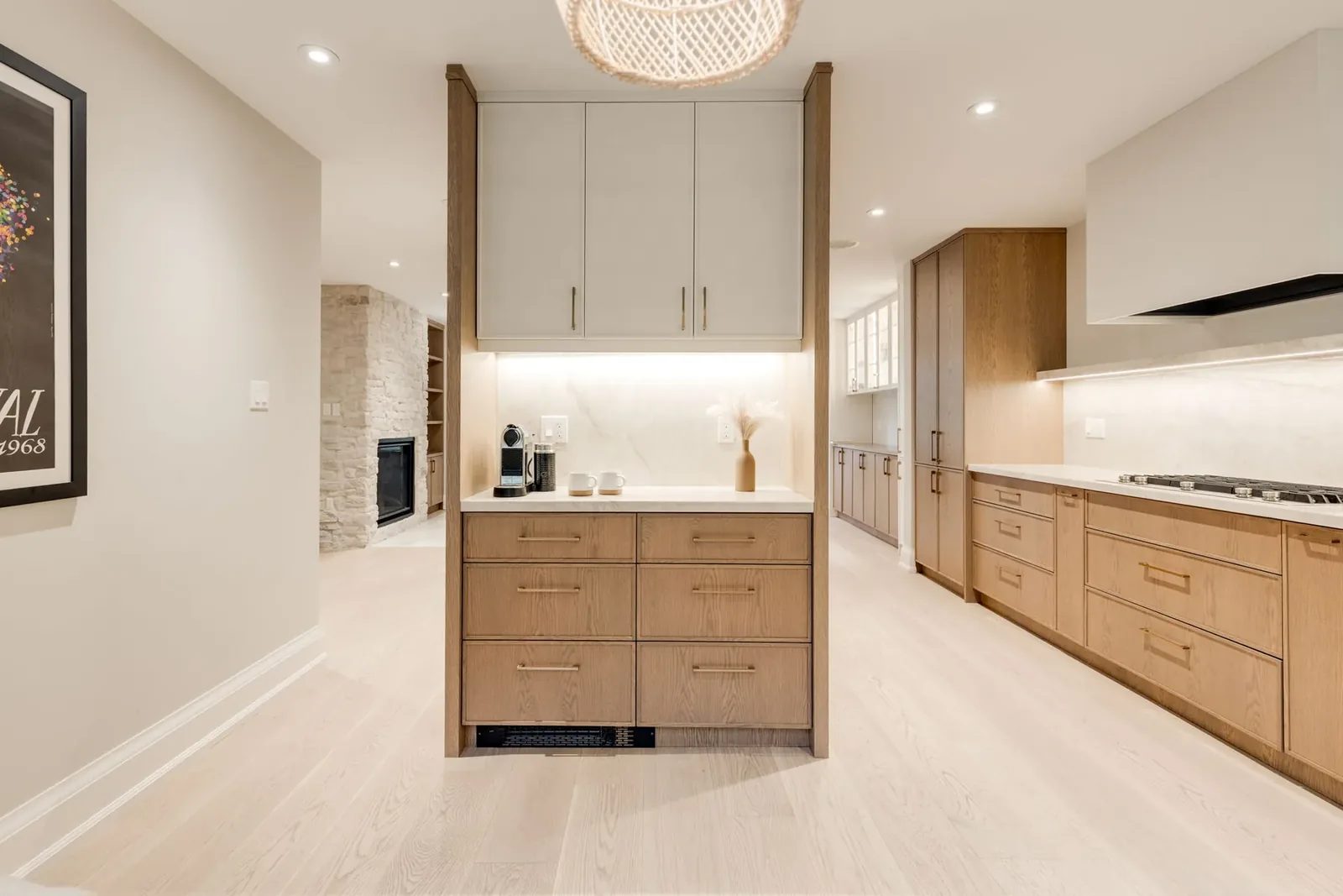White oak servery with its coffee station and stone counter beneath a woven pendant, opening to the kitchen run on one side and the whitewashed stone fireplace on the other.