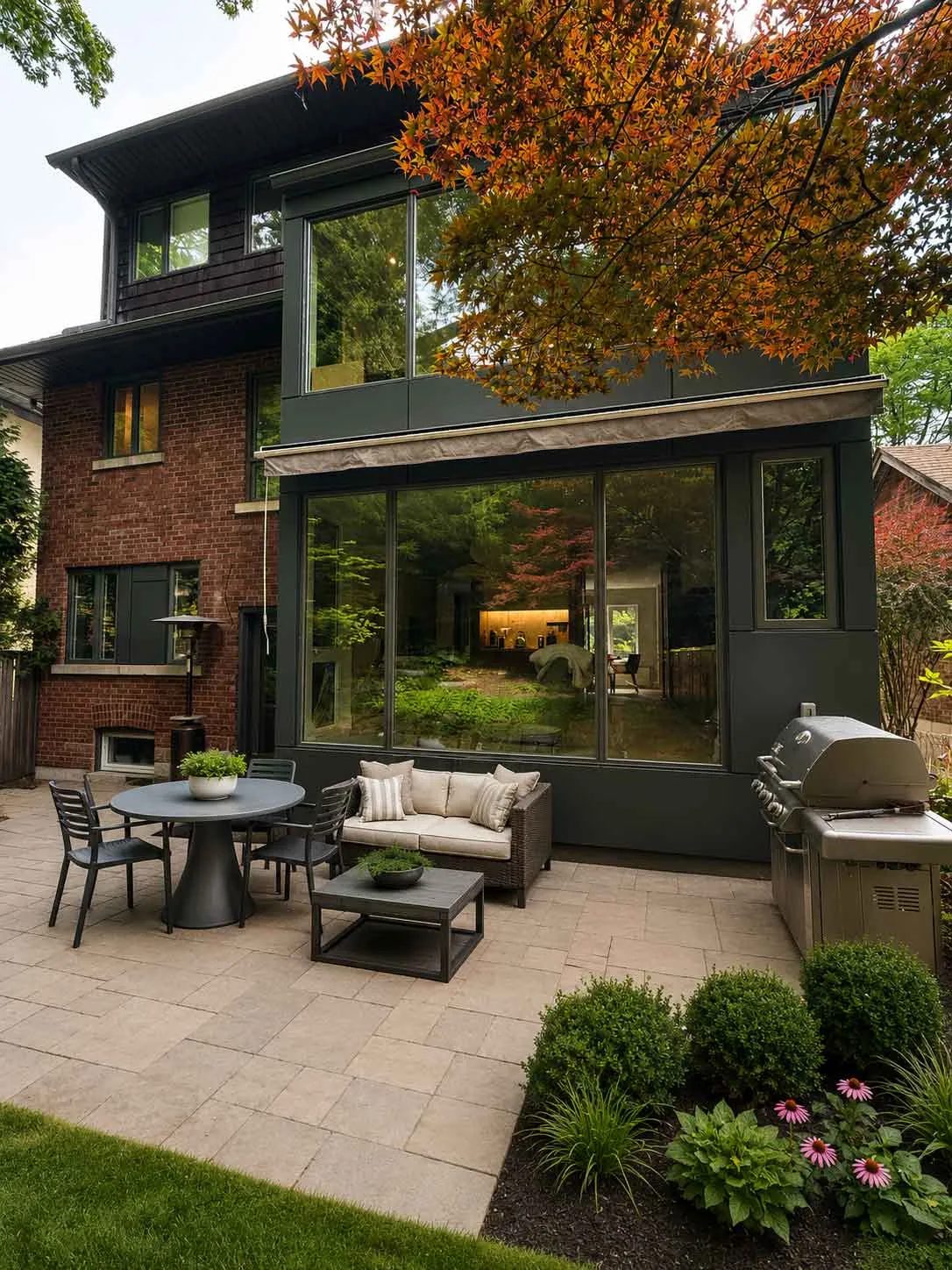 Rear of the house from the patio, the dark-clad glazed addition beside the original brick, with a dining table, woven lounge seating and planted borders on the pavers.