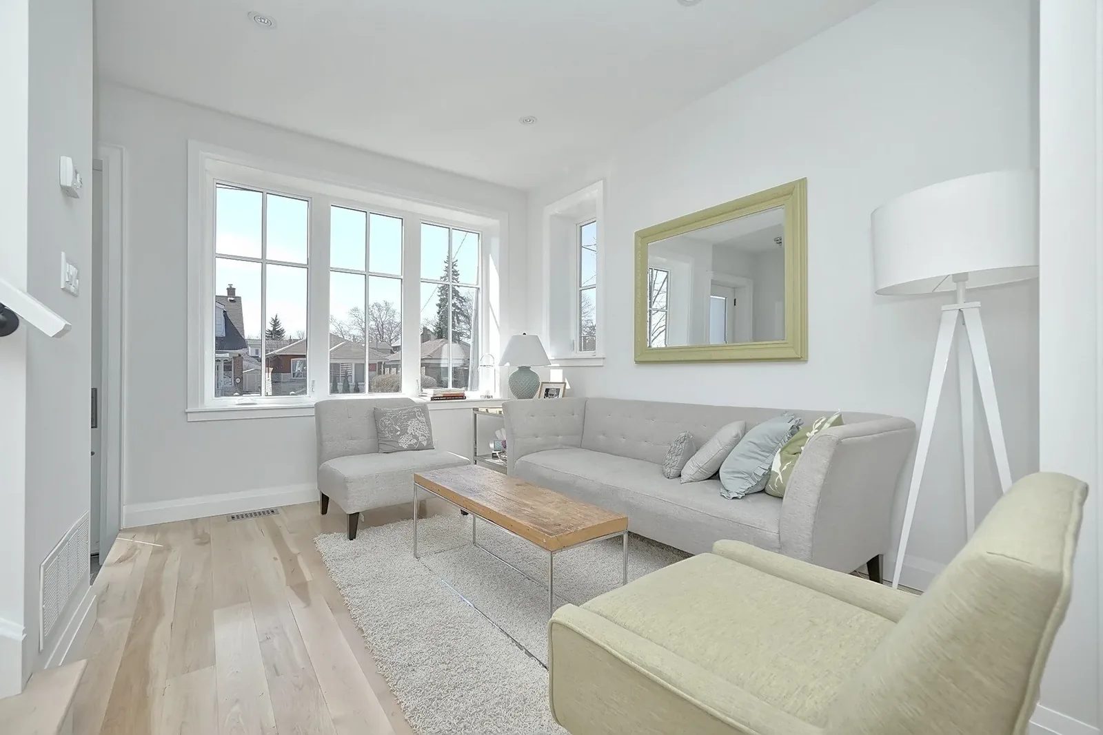 Bright Toronto living room with pale hardwood floors, large front windows, grey seating and warm white walls.