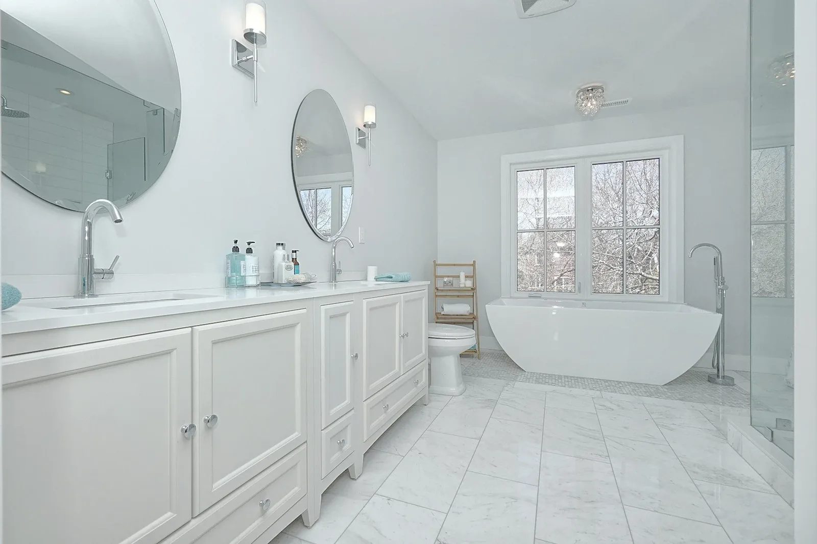 Bright white bathroom renovation with a double vanity, freestanding tub, glass shower, and marble tile floor.