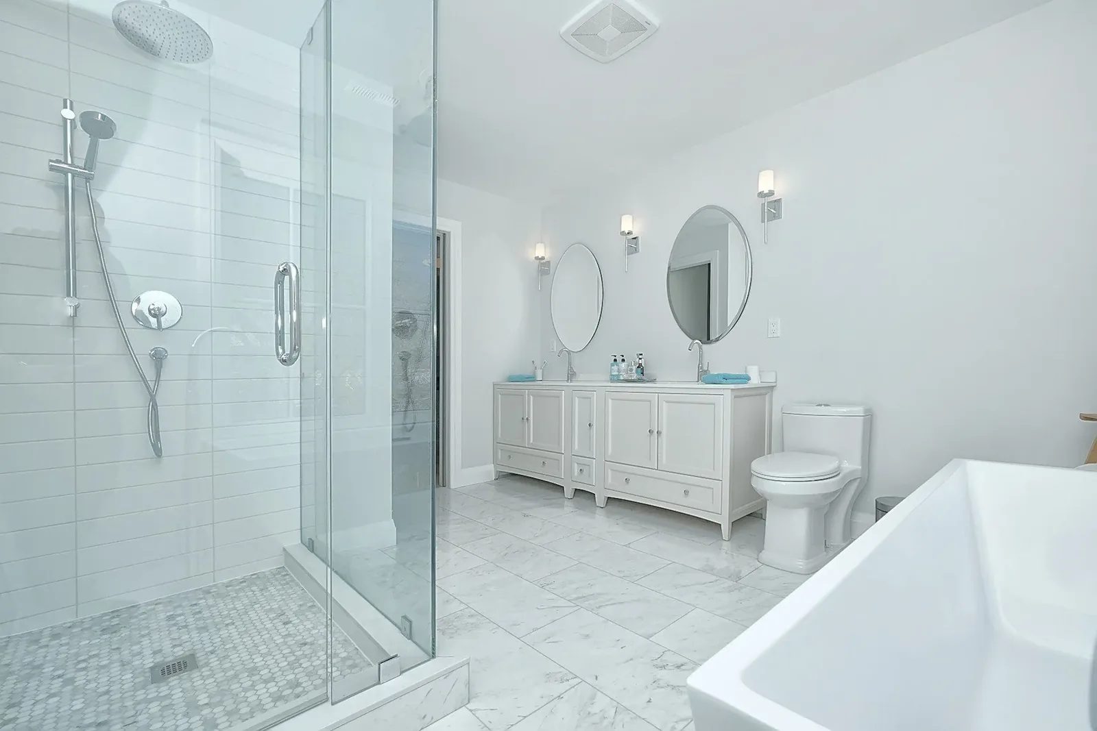 Bright white renovated bathroom with glass shower, double vanity, round mirrors and freestanding tub.