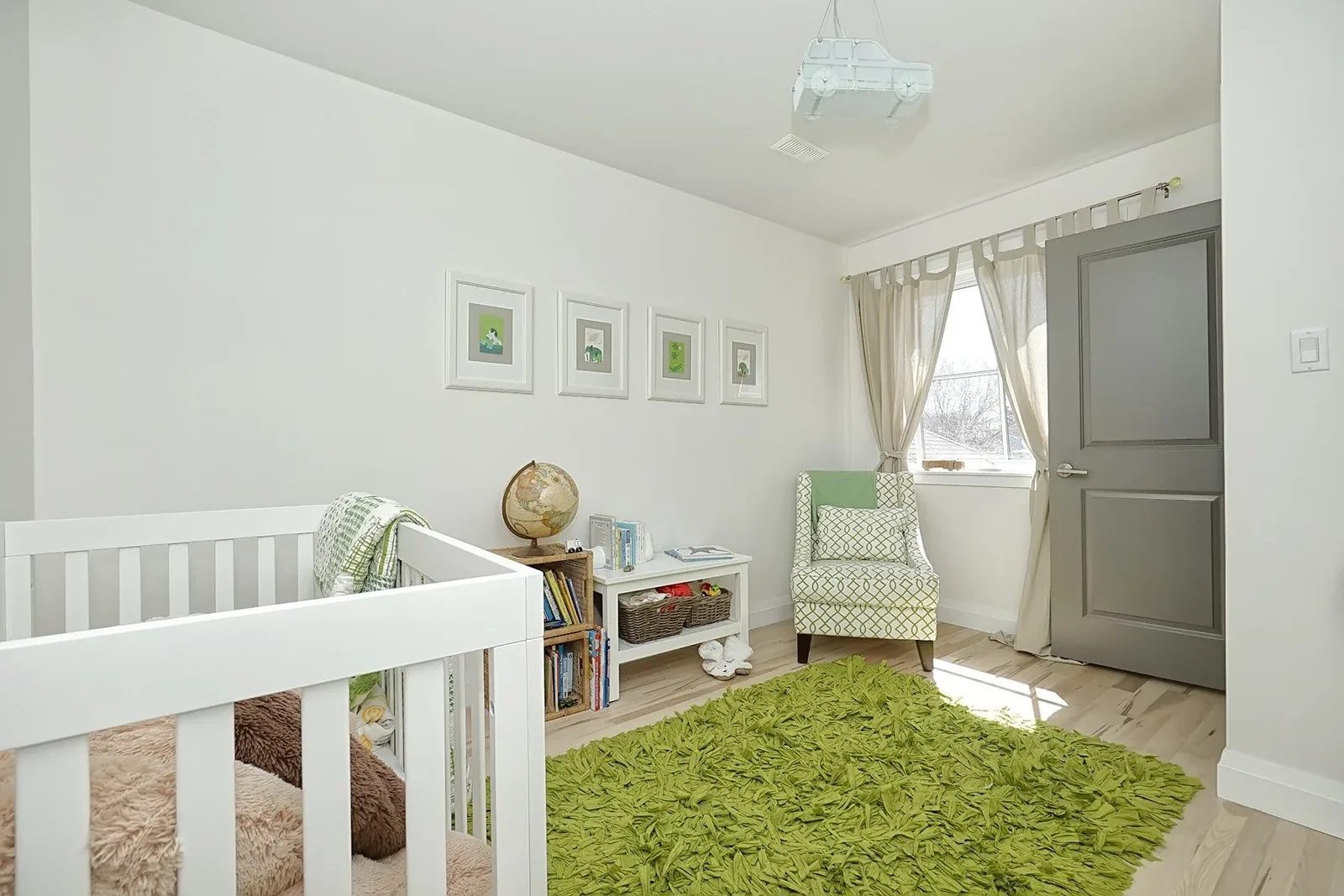 Bright nursery with white walls, light wood flooring, green rug, crib, armchair and grey door.