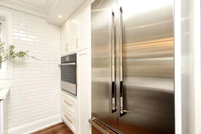 Stainless steel panelled refrigerator beside a built-in wall oven and white subway tile, Timeless Elegance project