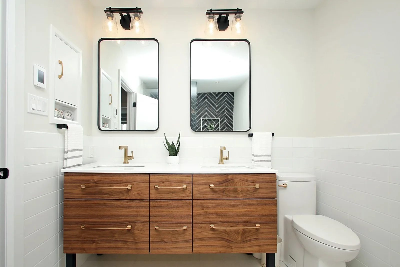 Bright bathroom with a walnut vanity, black mirrors, brass fixtures, and white tile walls.