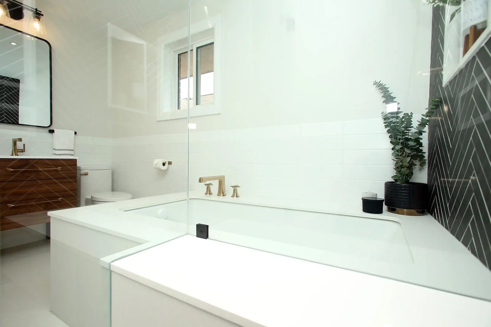 Bright white bathroom with a built-in tub, brass fixtures, dark herringbone tile and a wood vanity.