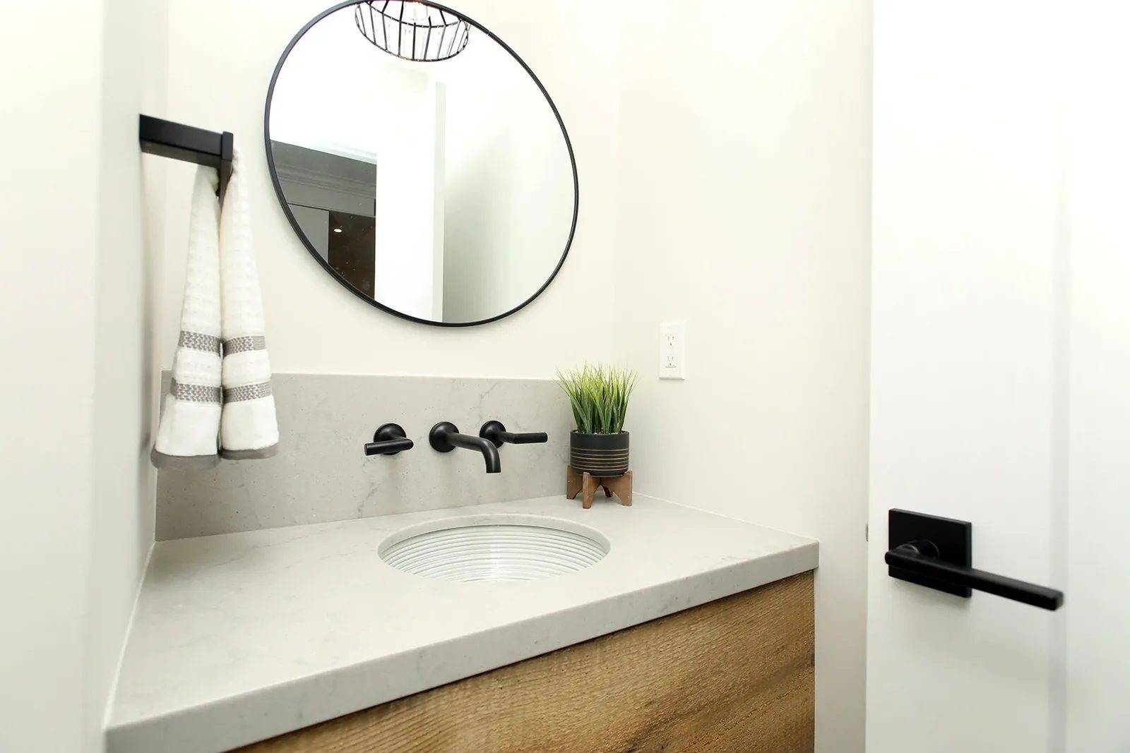 Bright powder room vanity with a round mirror, black wall taps, white stone counter, and warm oak cabinet.