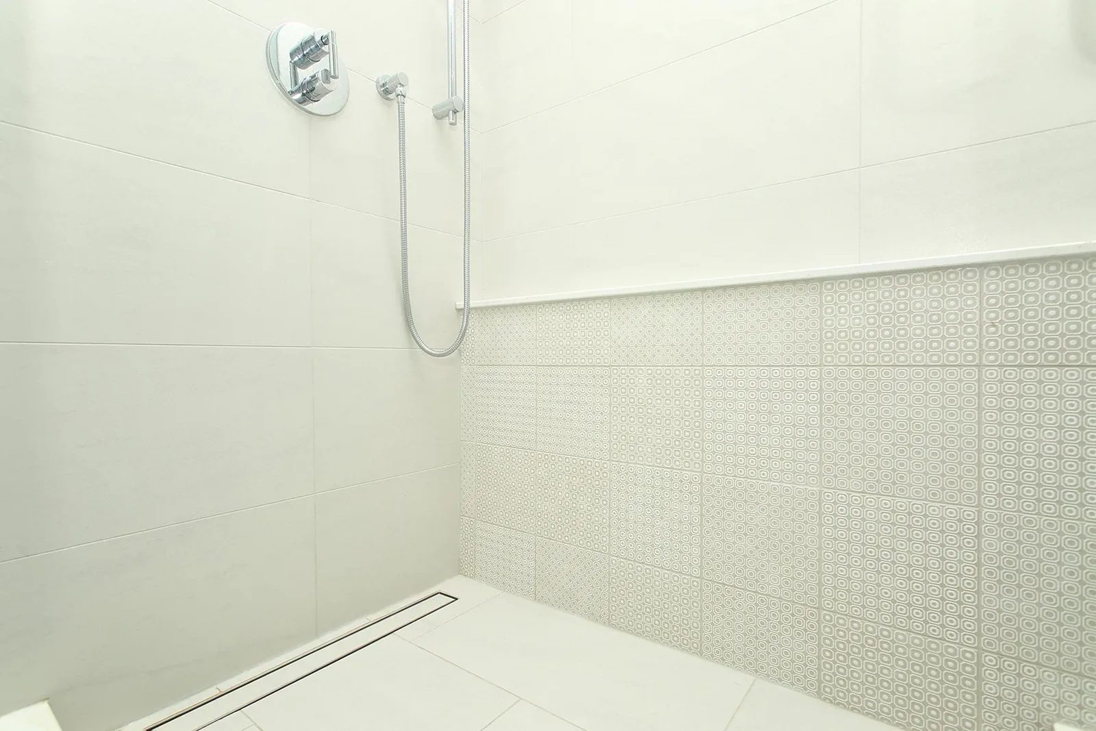 Warm white walk-in shower with patterned accent tile, chrome handheld fixture and a linear drain.