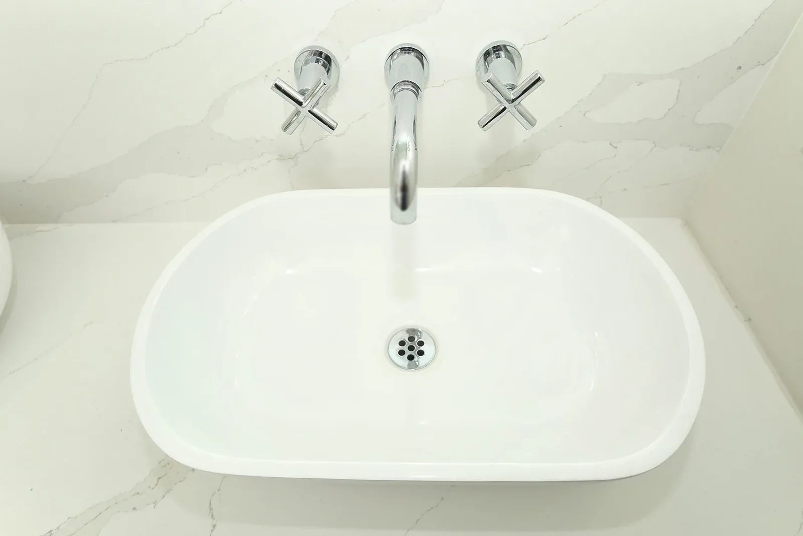 White bathroom sink with wall mounted chrome faucet, cross handles, and pale marble look quartz surround.