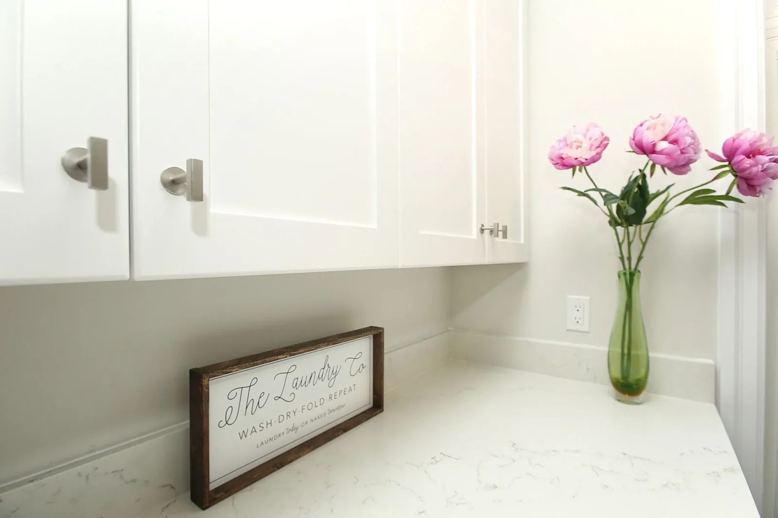 Warm white laundry room detail with shaker cabinets, quartz counter and pink peonies in a green vase.