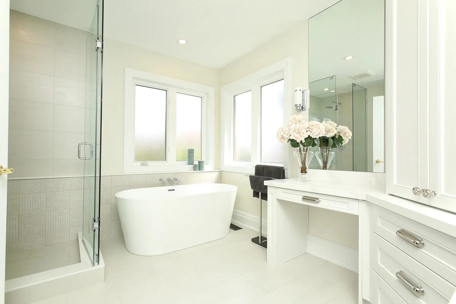 Bright white bathroom with a freestanding tub, glass shower, and built-in vanity beside frosted windows.