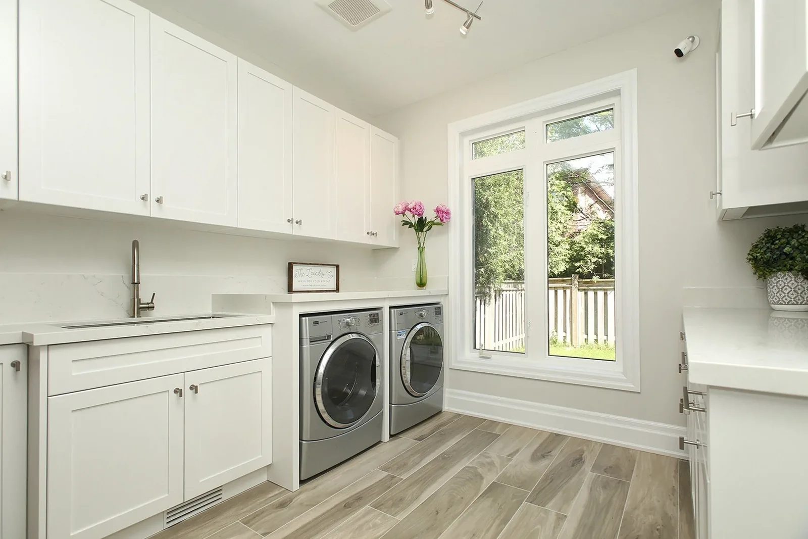 Bright white laundry room with quartz counters, shaker cabinets, and a large rear window.