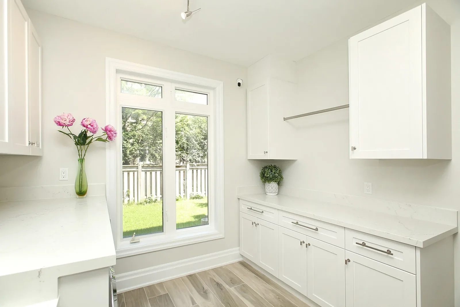 Bright white laundry room with shaker cabinets, quartz counters and a large window overlooking the yard.