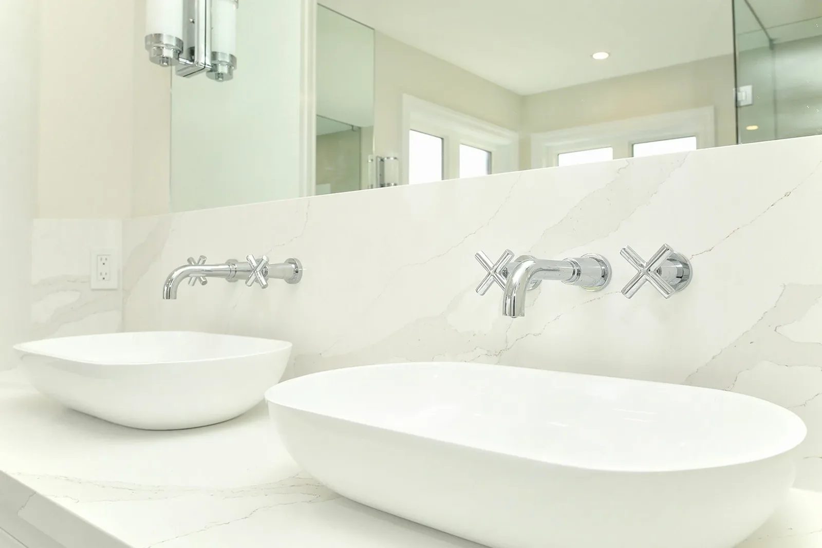 Bright bathroom vanity with two white vessel sinks, chrome wall taps, and a pale stone backsplash.
