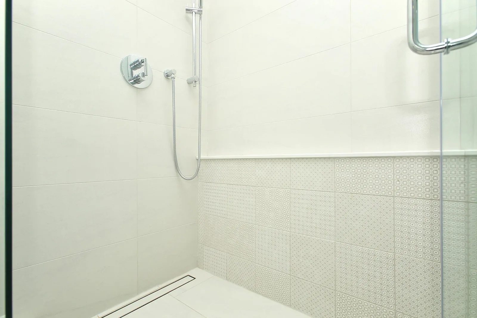 Bright walk-in shower with white tile, patterned accent walls, chrome fittings, and a linear drain.