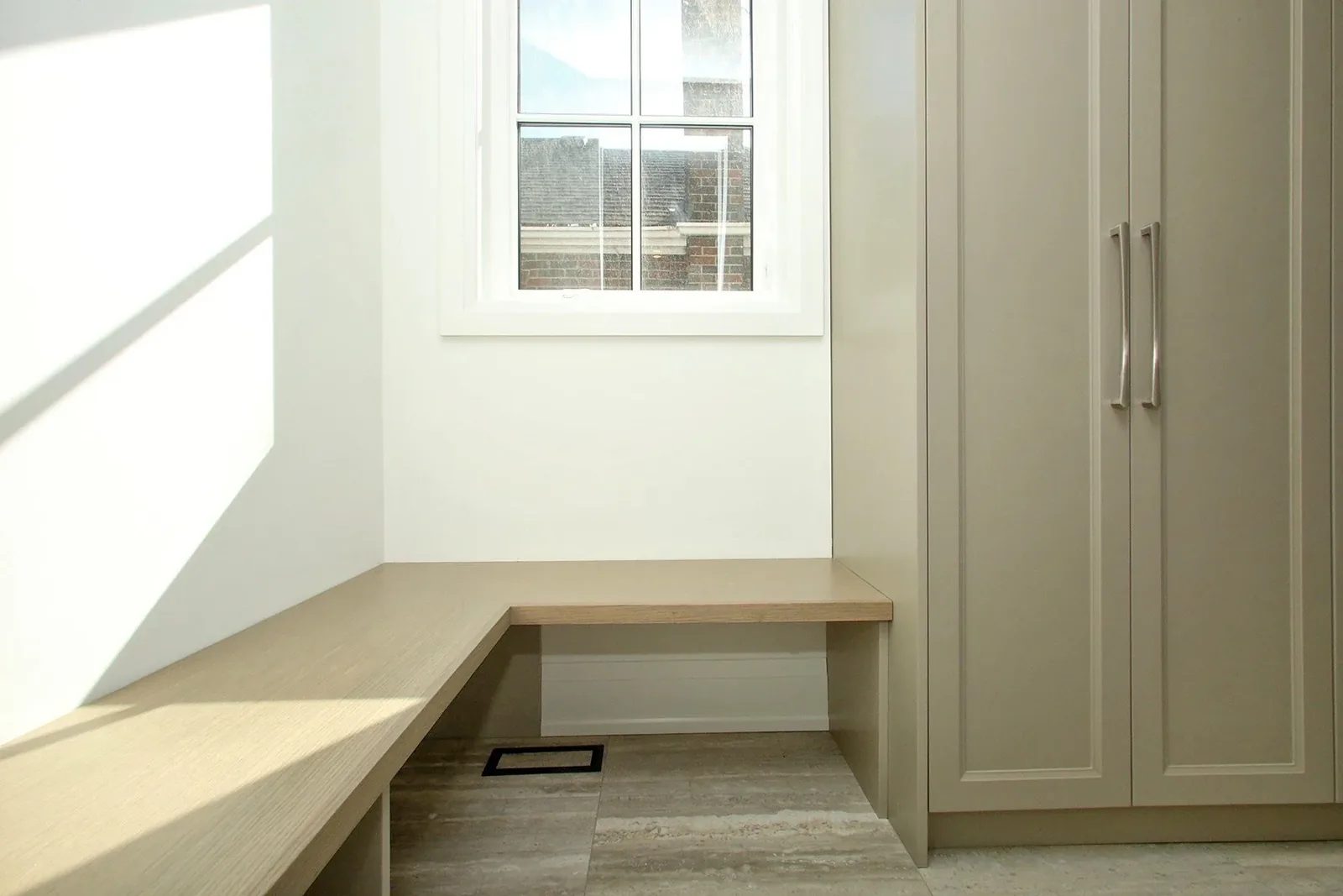Built in L shaped oak bench under a window beside warm grey storage cabinets in a mudroom.