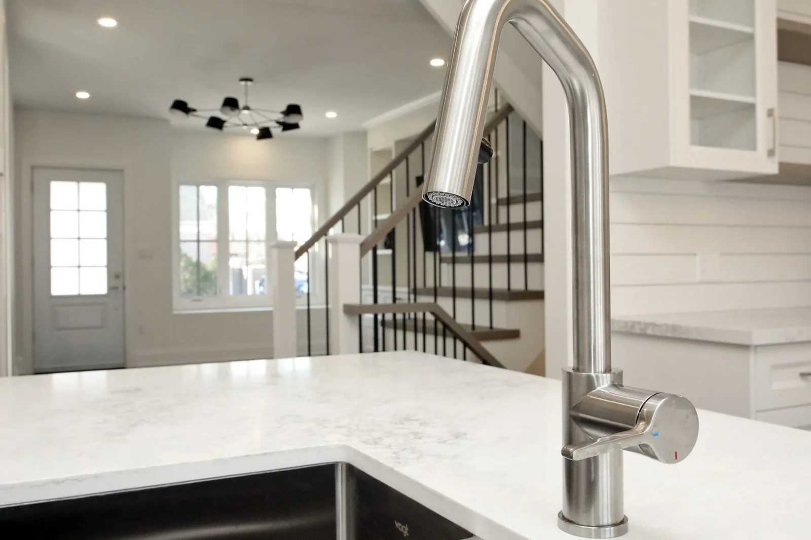 Brushed stainless gooseneck pull down kitchen faucet on a marble look quartz island with a staircase behind.