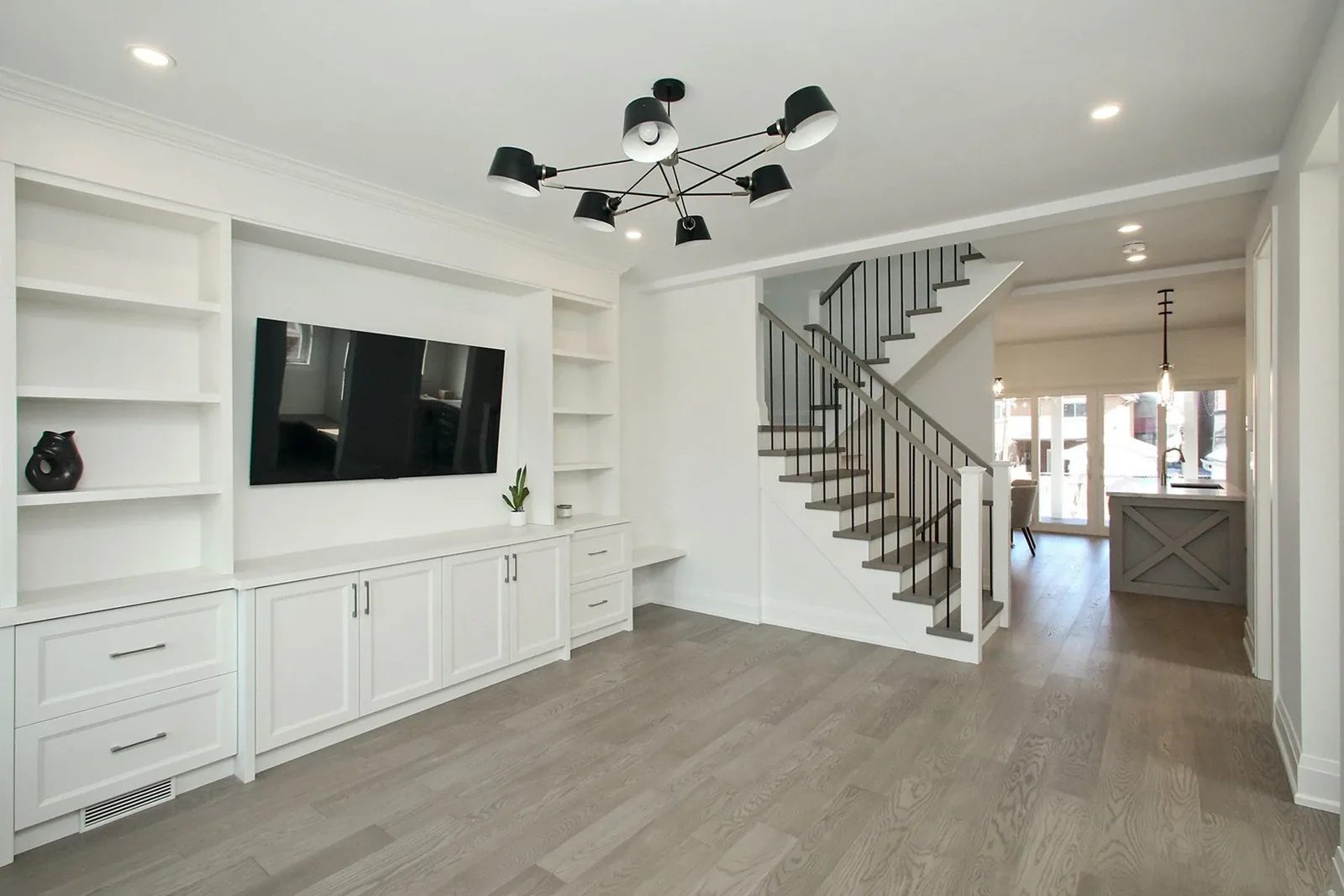 Open-concept main floor with white built-in media wall, modern black chandelier, and grey hardwood flooring.