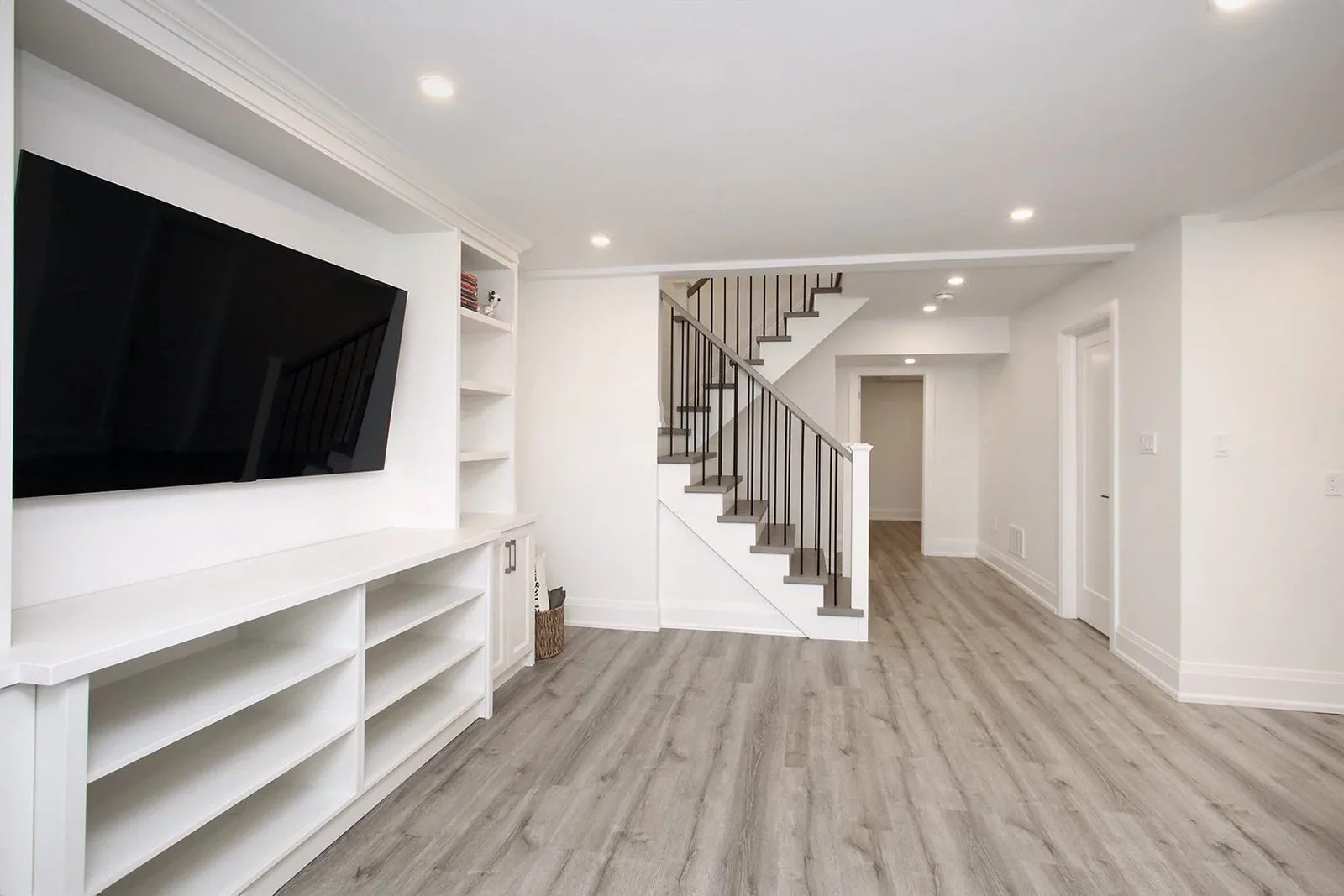 Finished basement recreation room with white built-in media unit, mounted television, and grey vinyl plank flooring.