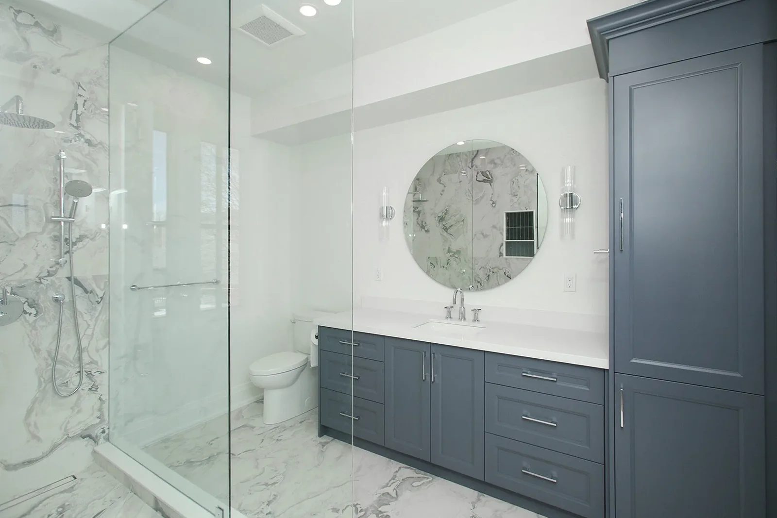 Primary ensuite with book-matched marble walls walk-in glass shower slate blue vanity and round mirror.