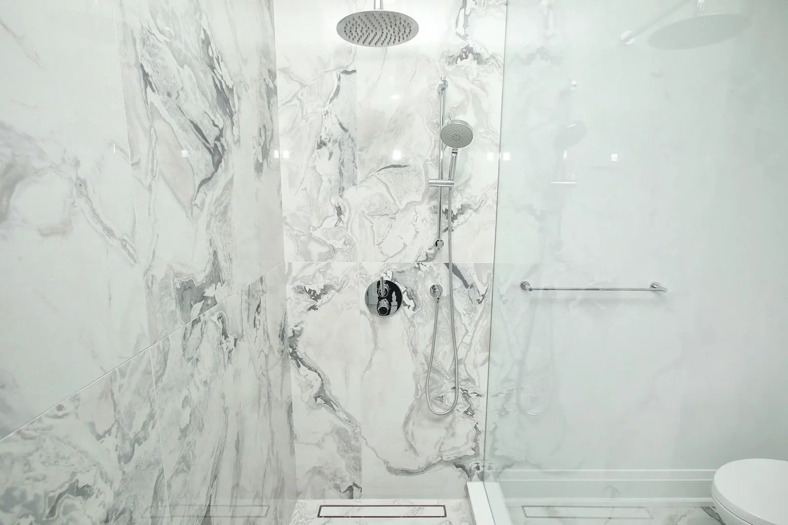 Close-up of walk-in shower with book-matched marble slab walls rain head and chrome handheld fixture.