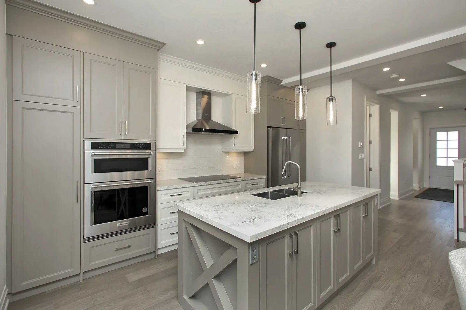Warm grey transitional kitchen with an X panel island, wall ovens, and a chimney hood over the cooktop.