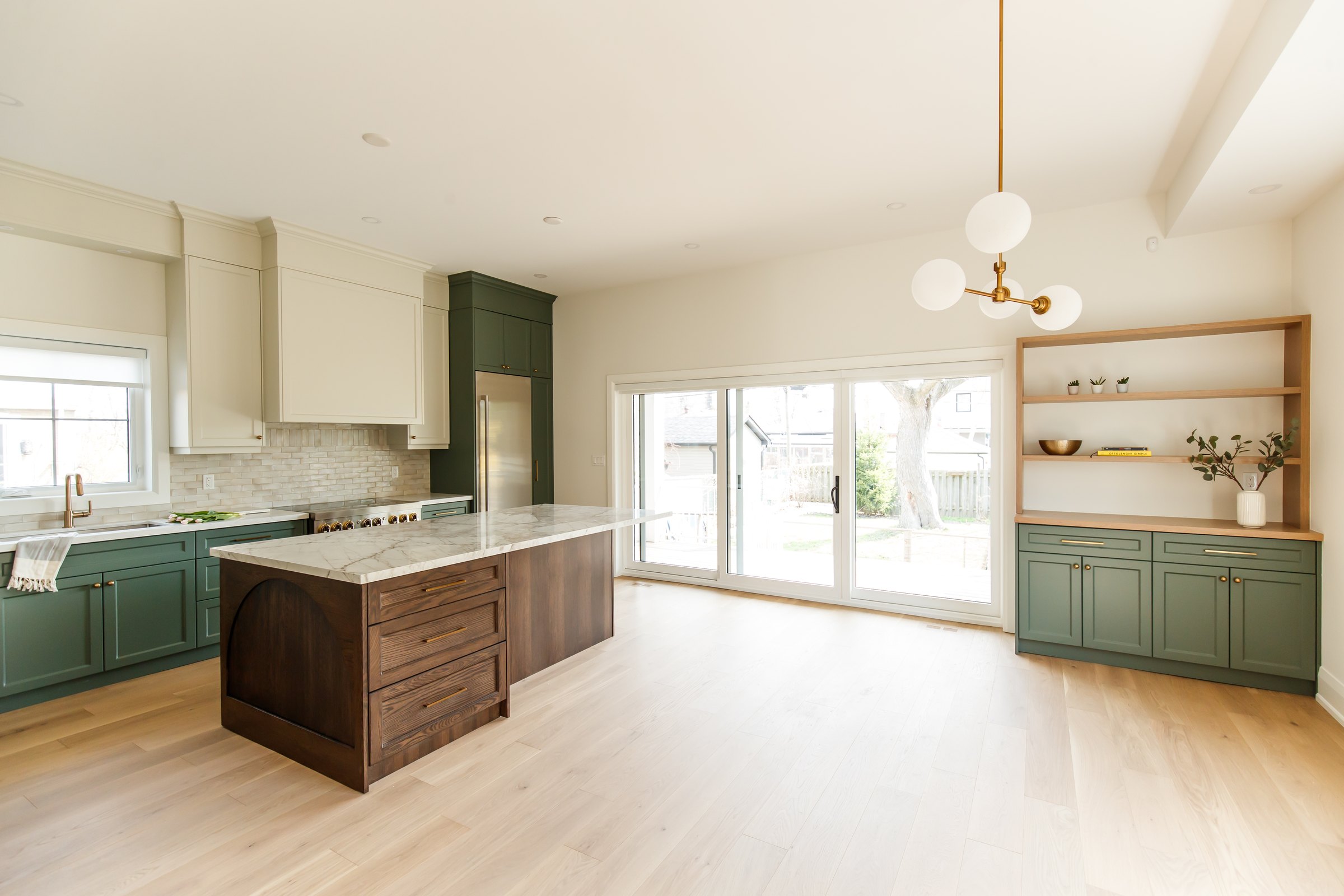 Inside a finished Waterfront kitchen in Etobicoke: sage-green and cream cabinetry runs along the wall to a marble-topped walnut island, with a tiled backsplash, a gas range, and glass doors opening to the back garden.
