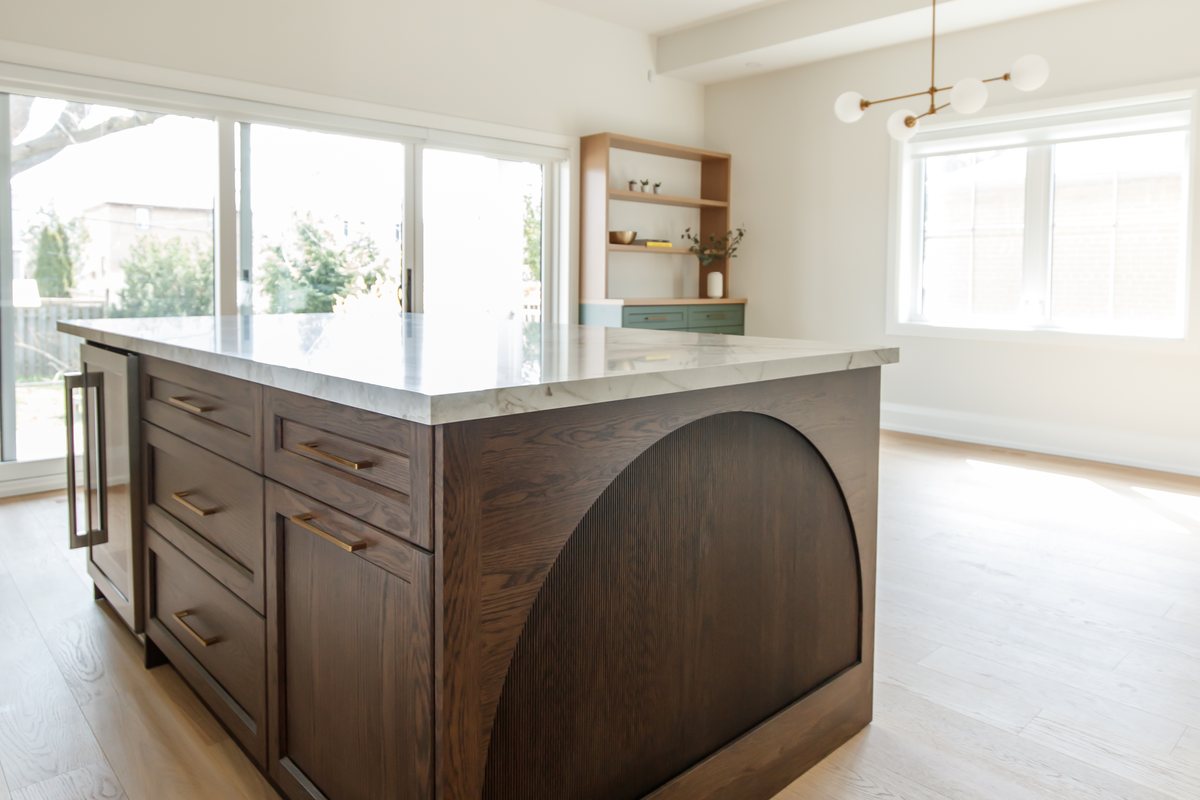 A close view of Waterfront kitchen millwork: a custom walnut island with an arched fluted end panel, brass drawer pulls, and a marble waterfall edge on light oak floors.
