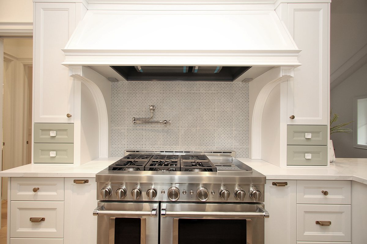 A Waterfront kitchen range wall: a professional dual-oven range under a custom hood, framed by a patterned tile backsplash and sage-green spice drawers.